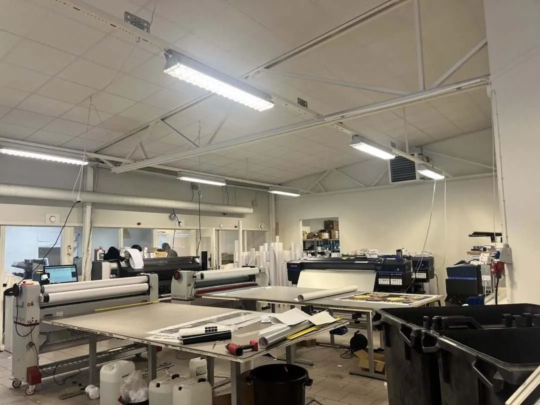 A spacious printing workshop with various machines and equipment. The room is well-lit with fluorescent lights and has a clean, organized layout. There are larg